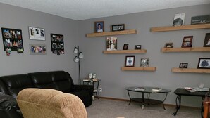 A living room with a brown armchair, a black leather couch, a glass coffee table, and a small round table with books and a cup. Shelves with framed photos and a lamp are on the wall. - Private condo sleeps up to 4 with private hot tub (Lincoln)