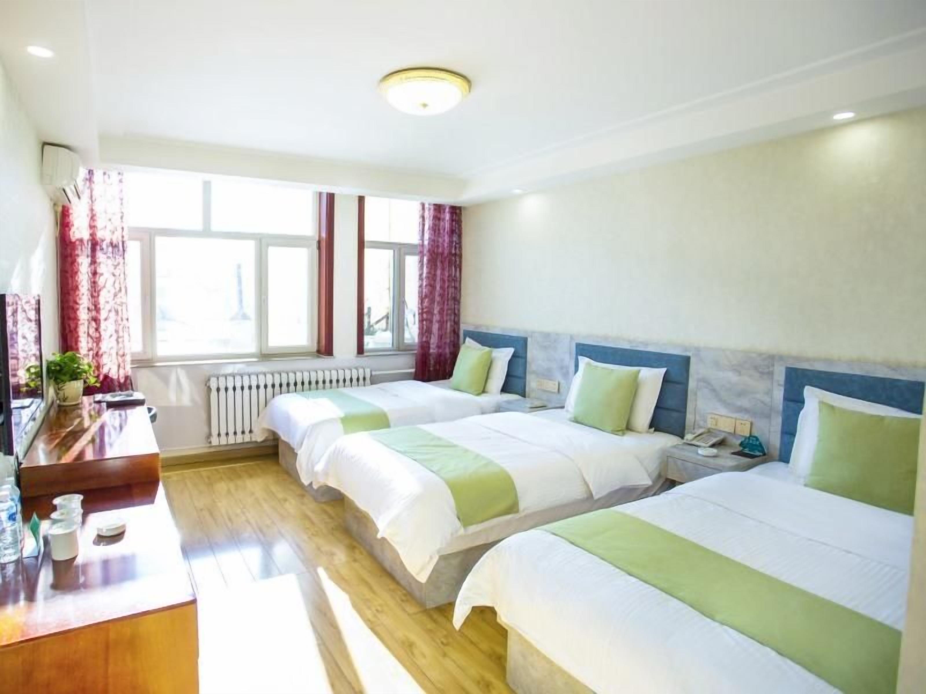 A hotel room with two beds, a wooden desk, a radiator, and a window with curtains.
