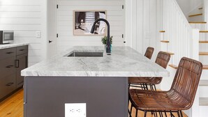 Una cocina moderna con una isla de mármol, lámparas colgantes negras, sillas de mimbre y una escalera.