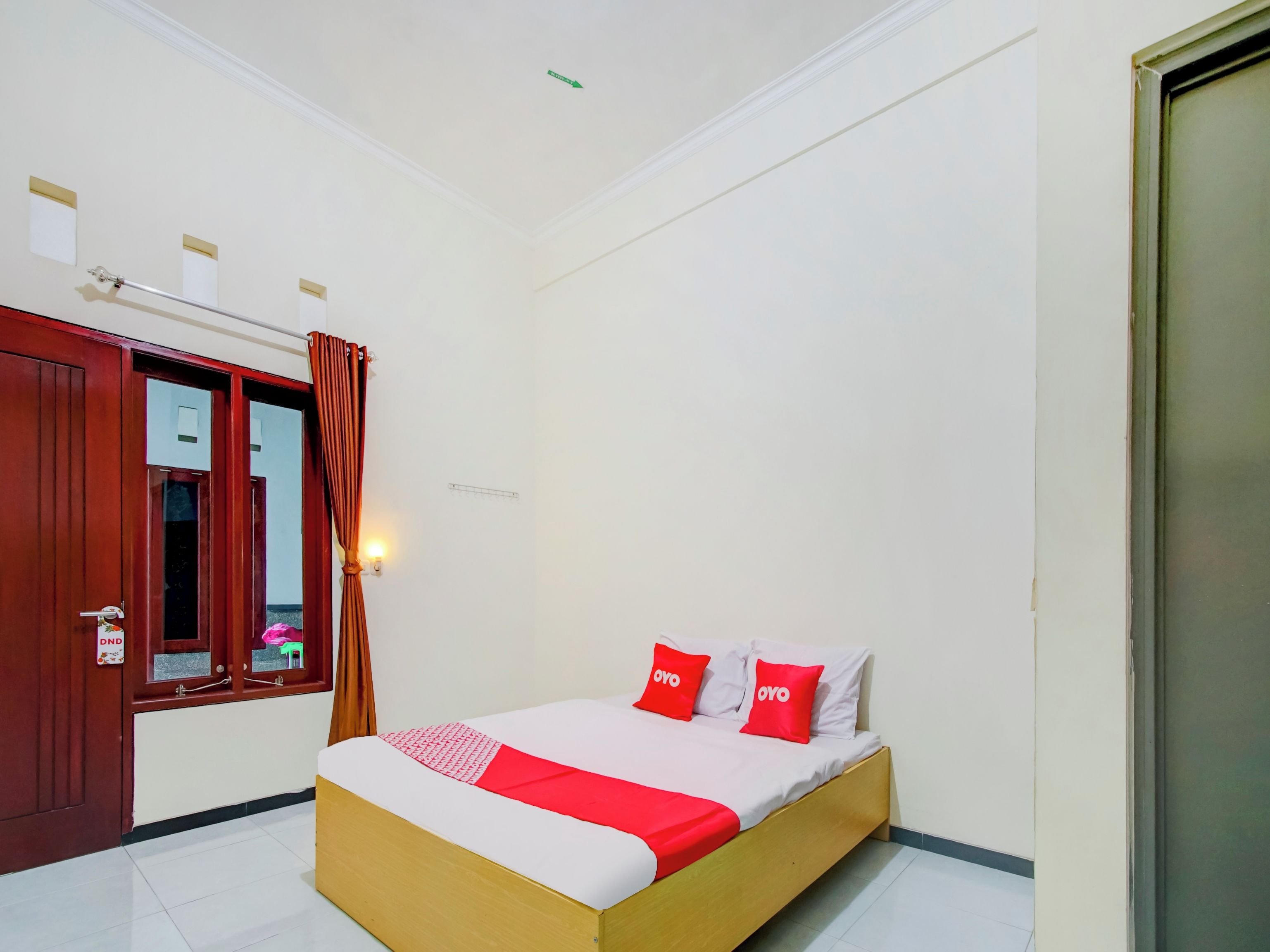 A bedroom with a wooden bed, red and white bedding, a mirror, a door with a sign, and a wall-mounted light fixture.