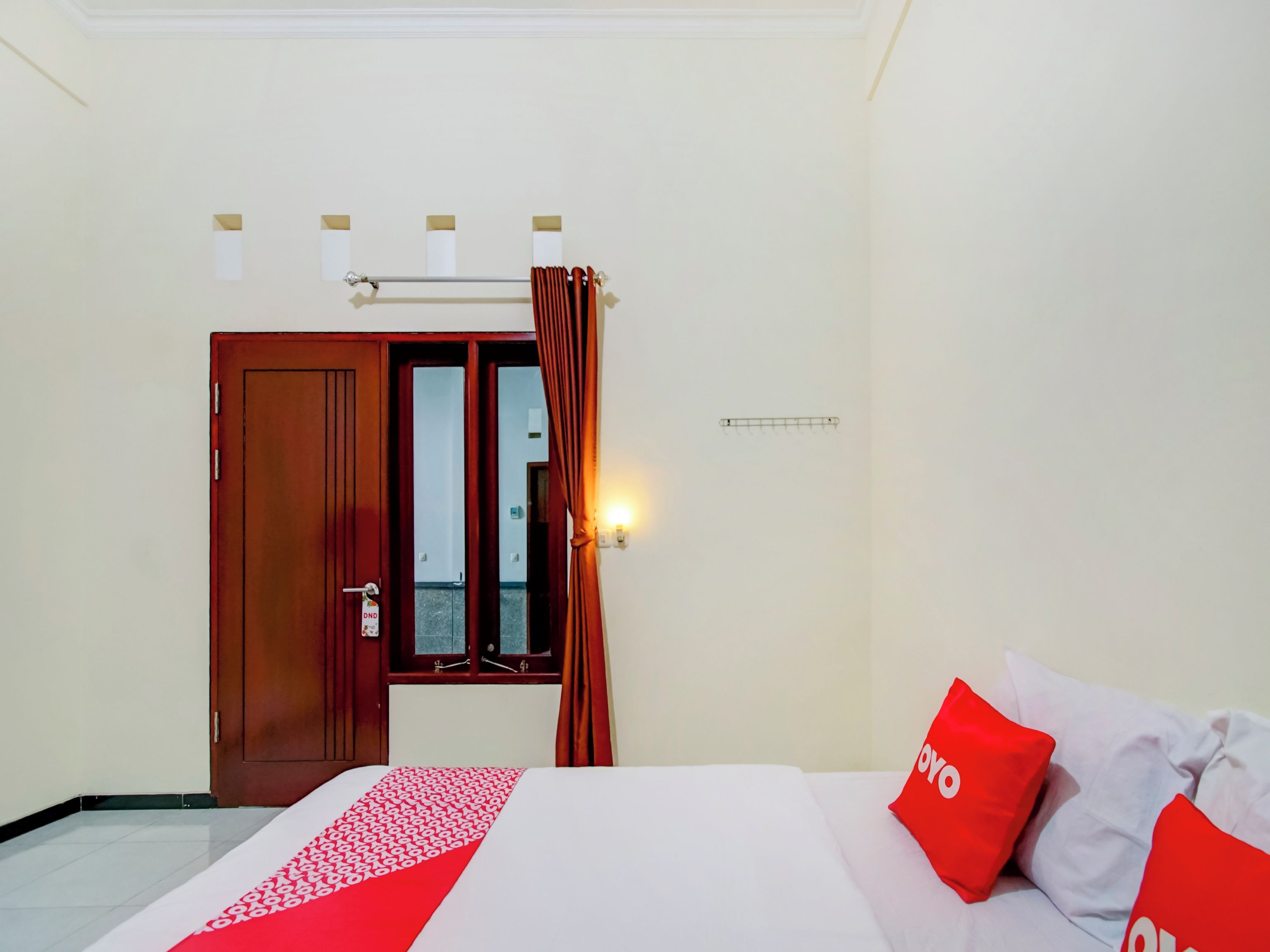Habitació d'hotel amb un llit, una porta de fusta, una finestra amb cortines i una làmpada subjecta a la paret.