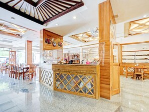 Un hall d’hôtel spacieux, doté d’un comptoir de réception central en bois, de coins salon et d’un plafond haut avec des poutres apparentes.