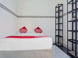 Un lit bien fait, recouvert d’une couvre-lit blanche et rouge, avec deux oreillers rouges ornés de motifs blancs, et une étagère métallique noire adossée au mur.