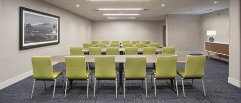 A conference room with a long table and green chairs, a framed cityscape picture on the wall, and a lamp on a side table.