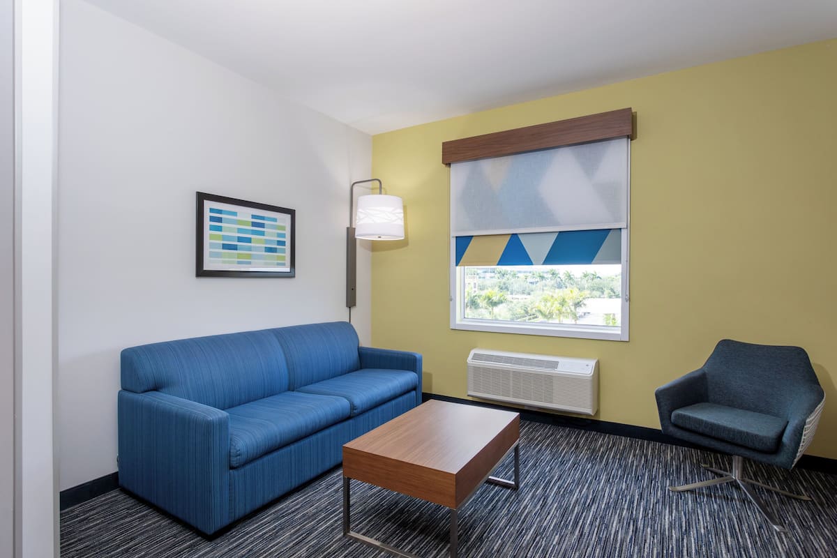 A room with a blue sofa, a wooden coffee table, a blue armchair, a framed picture on the wall, and a window with blinds.