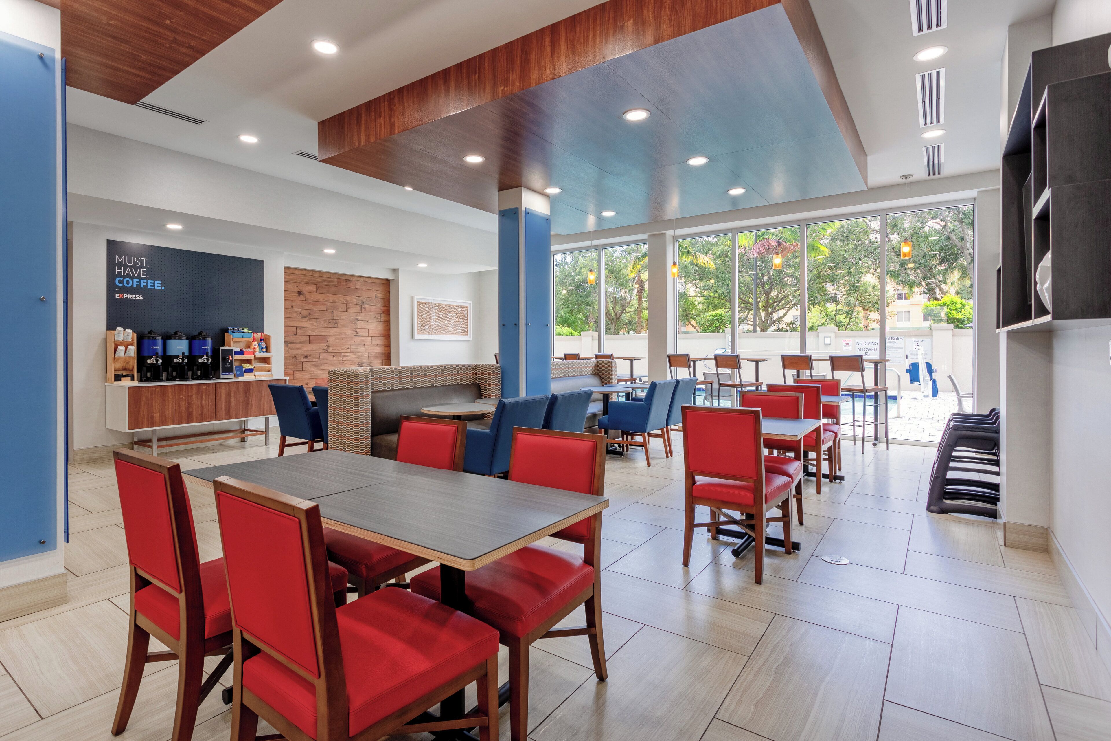 A modern coffee shop with a wooden ceiling, large windows, and red and blue seating areas.