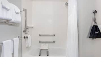 Une salle de bain moderne avec des carreaux blancs, une baignoire, une douche et une barre d’appui. Des serviettes sont suspendues à un porte-serviettes et un sac noir est accroché au mur.