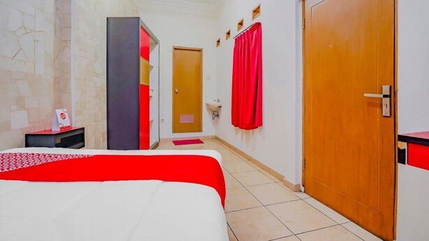 A hotel room with a bed, a wooden door, a bathroom with a mirror, and a red curtain. - OYO 90035 Merci Club (Bandung)
