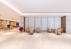A spacious lobby with a modern design, featuring a seating area with wooden chairs and a red accent chair, a large wall with vertical blinds, and a well-lit ceiling with recessed lighting.