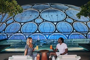 Two people sitting at a table with drinks in front of a modern architectural structure with a pool.