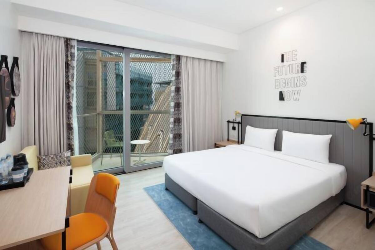 Moderni hotellihuone, jossa on suuri sänky, työpöytä, tuoli ja lasiovesta avautuva näkymä.