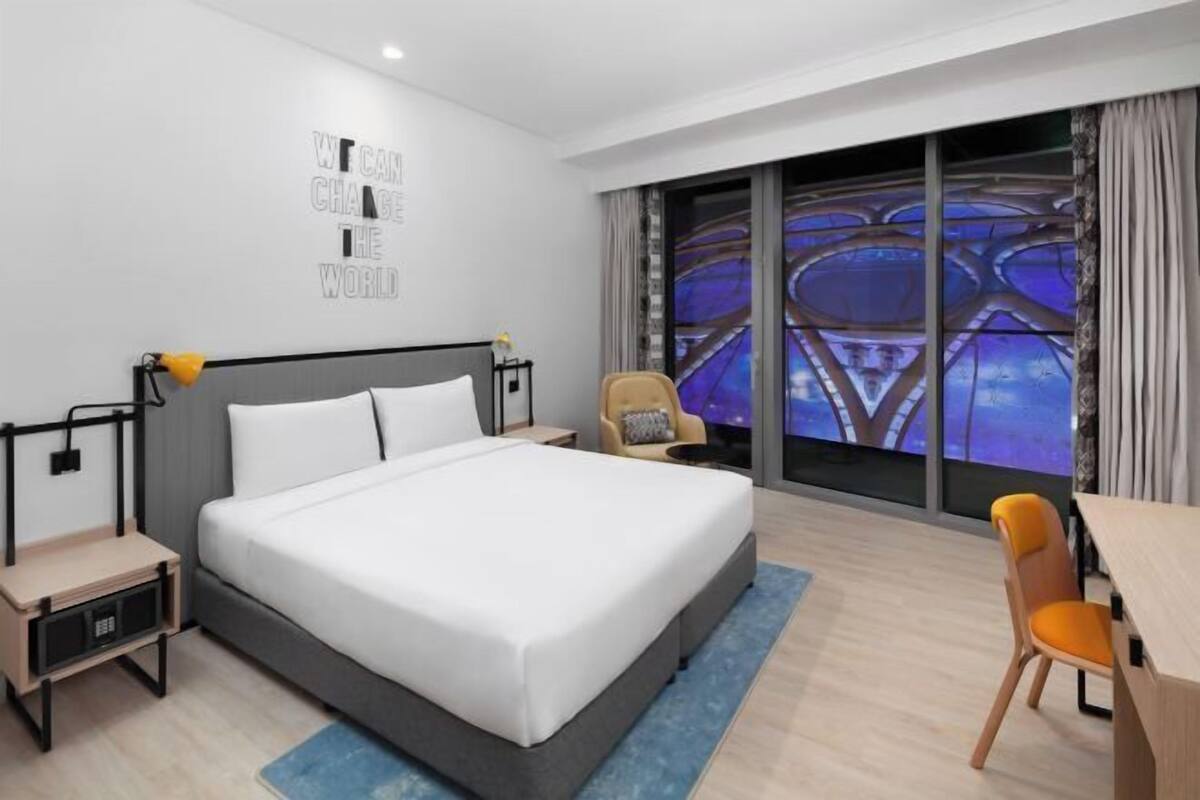 Moderni hotellihuone, jossa on suuri sänky, työpöytä, tuoli ja suuri ikkuna, josta avautuu näkymä kaupunkimaisemaan.