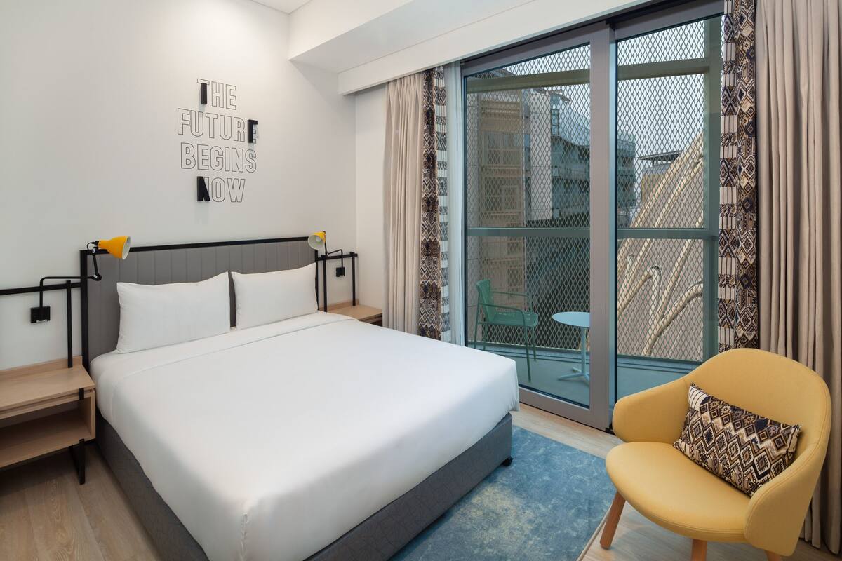Moderni hotellihuone, jossa on suuri sänky, pieni pöytä, tuoli ja liukuovista lasiovesta avautuva näkymä ulkoalueelle.