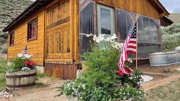 A wooden cabin with a number '9215' on the door, surrounded by desert vegetation and two American flags. - Ursa Miner - quiet, dark sky, 6 miles from Pitkin! (Parlin)