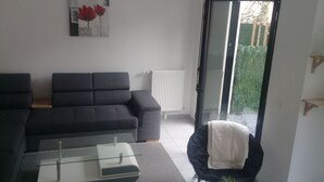 A living room with a dark sofa, a glass coffee table, a radiator, and a sliding glass door leading to a garden. - Hortillonne (AMIENS)