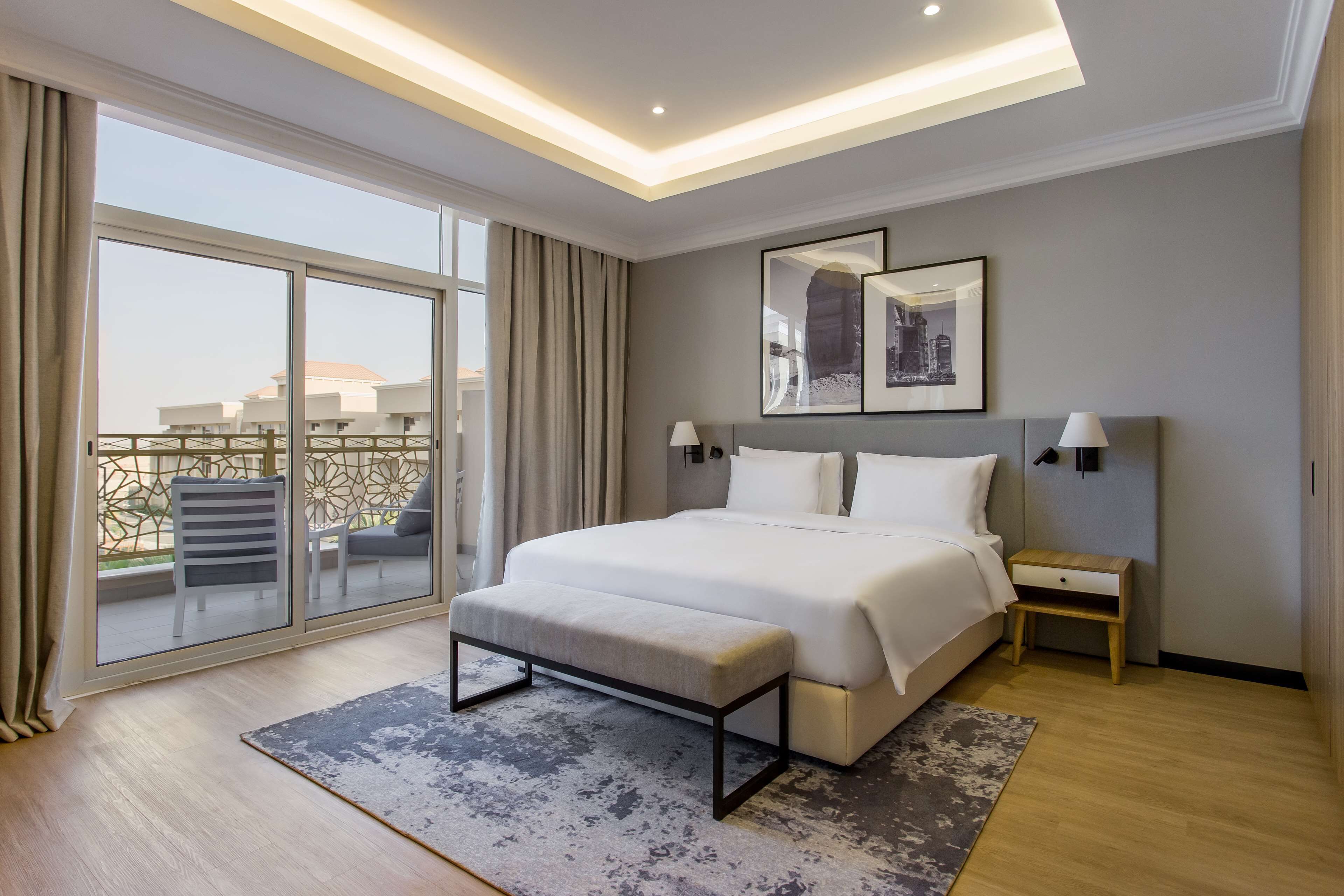 A modern hotel bedroom with a large bed, a bench, a nightstand, and a view of the city through large windows.