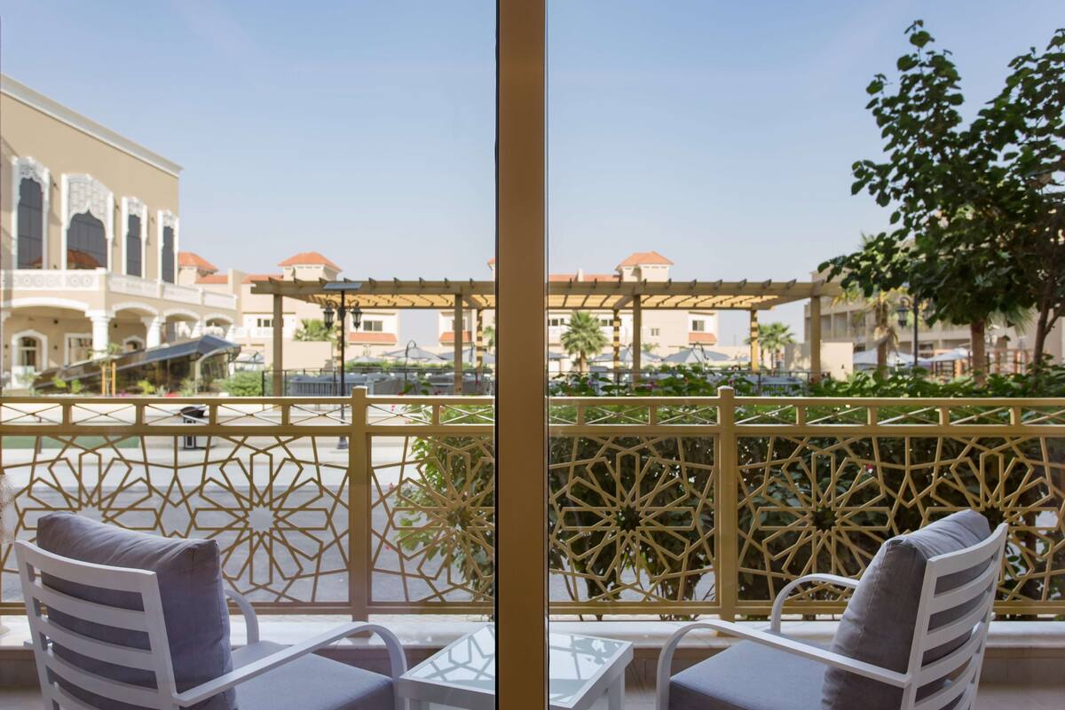 A balcony with white chairs and a table, overlooking a courtyard with a decorative fence and buildings in the background.