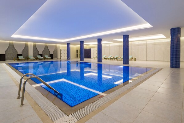 An indoor swimming pool with a blue tiled floor, a metal ladder, and lounge chairs along the sides.