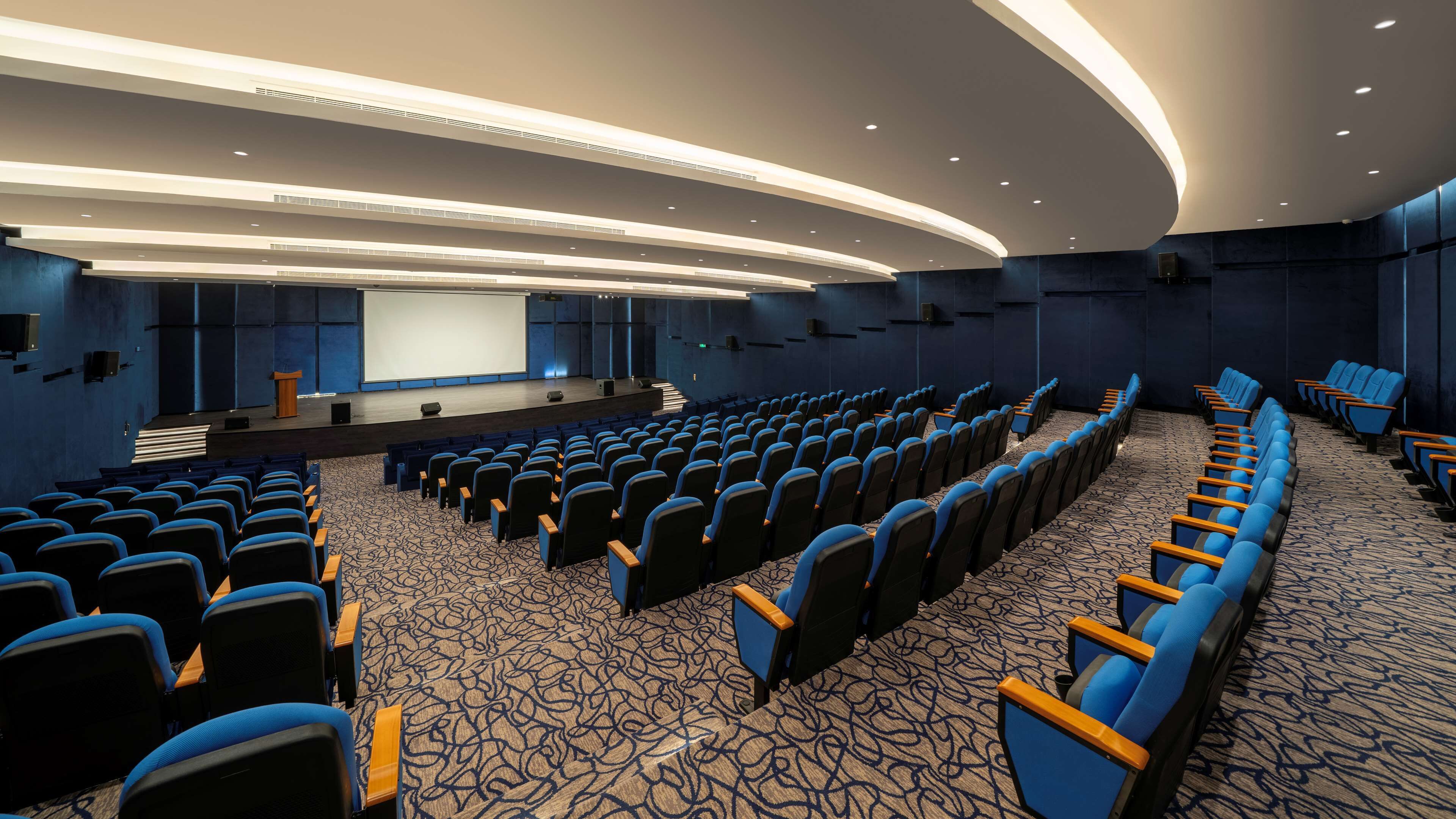 Une salle de conférence avec des rangées de chaises bleues, une scène équipée d’un projecteur et un sol recouvert de moquette.