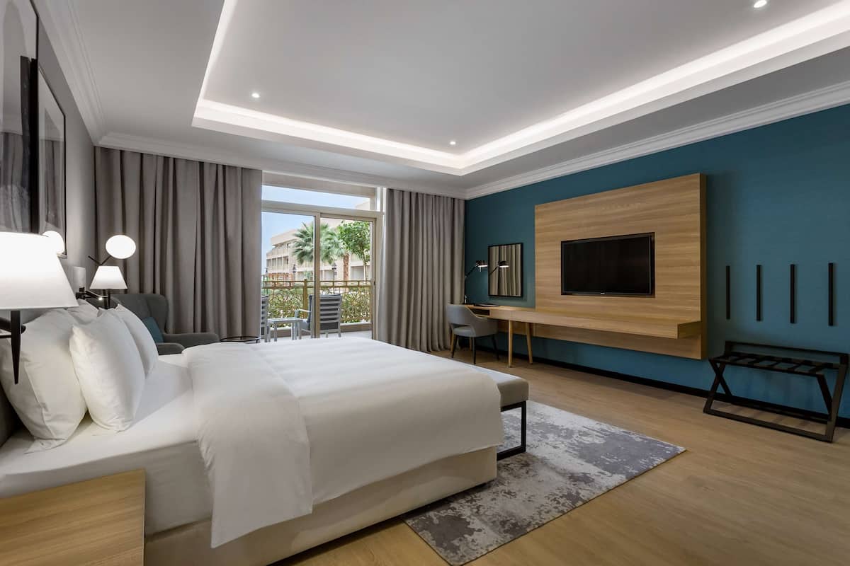 A modern hotel room with a large bed, a desk, a TV mounted on the wall, and a view of the outdoors.