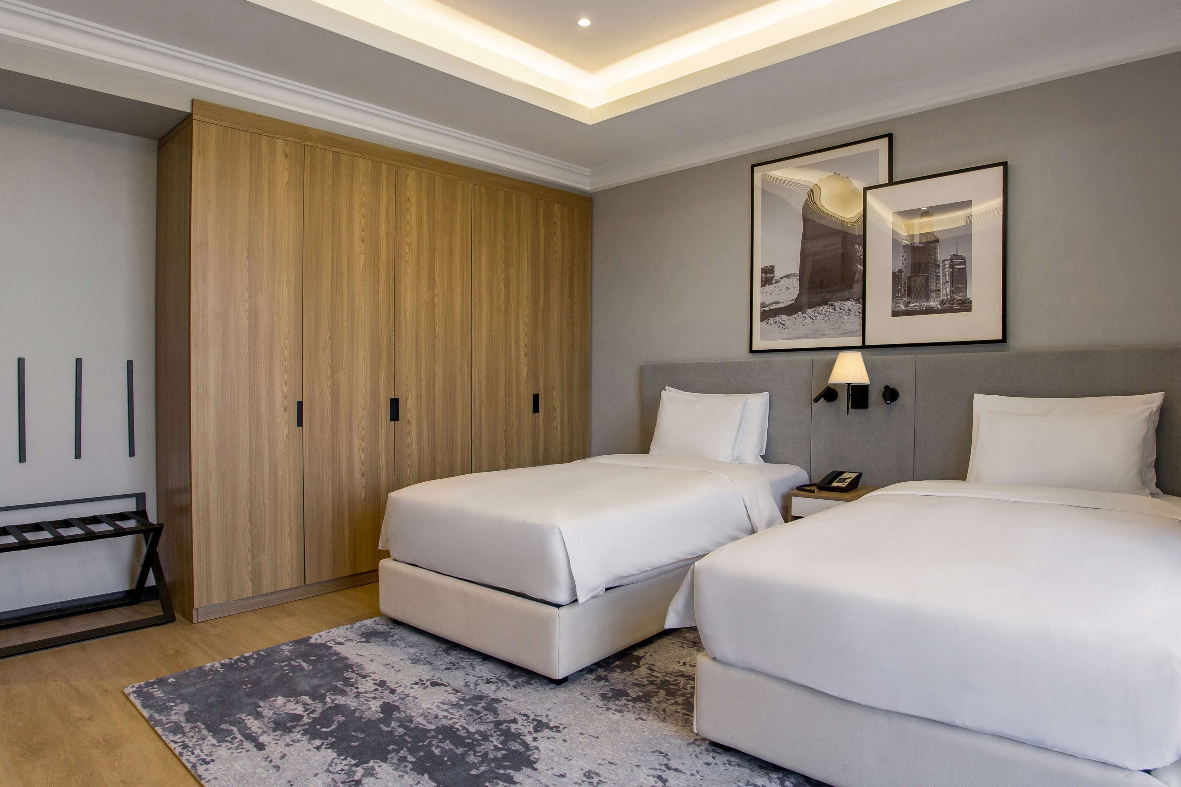 A hotel room with two beds, a wooden wardrobe, a grey rug, and two framed pictures on the wall.