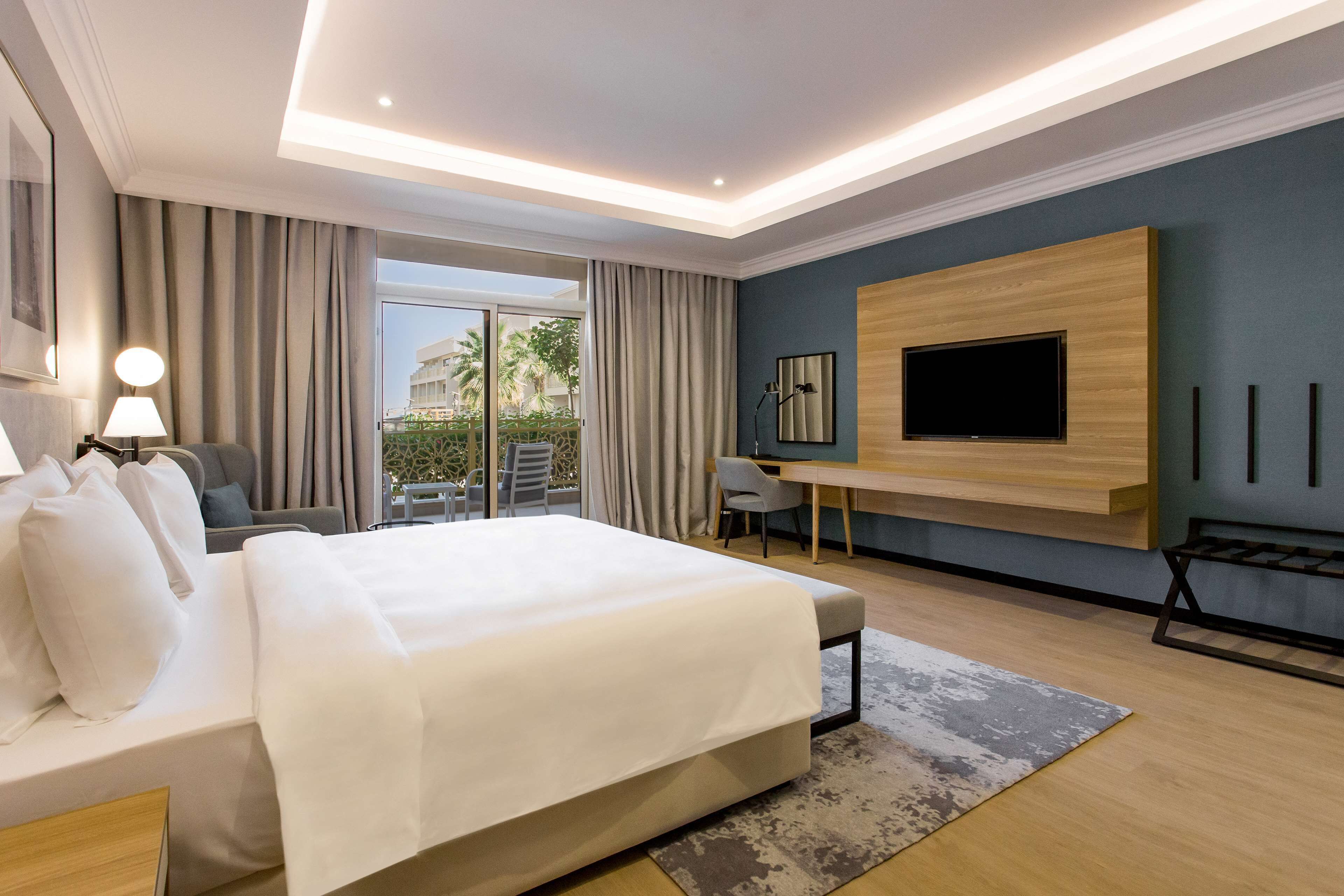 A modern hotel room with a large bed, a desk, a flat-screen TV mounted on the wall, and a balcony with outdoor seating.