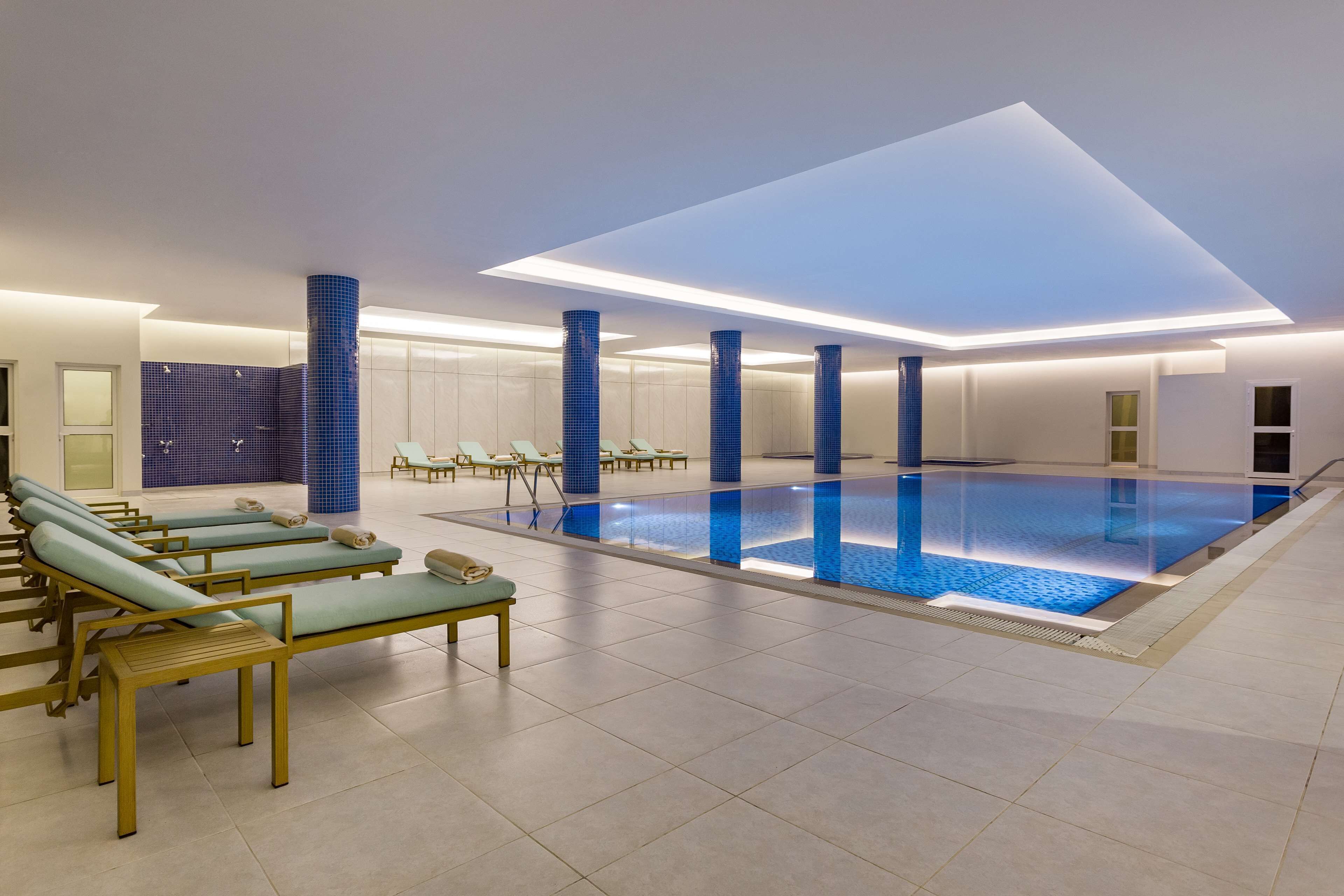 A modern indoor swimming pool with lounge chairs and a blue-tiled floor.