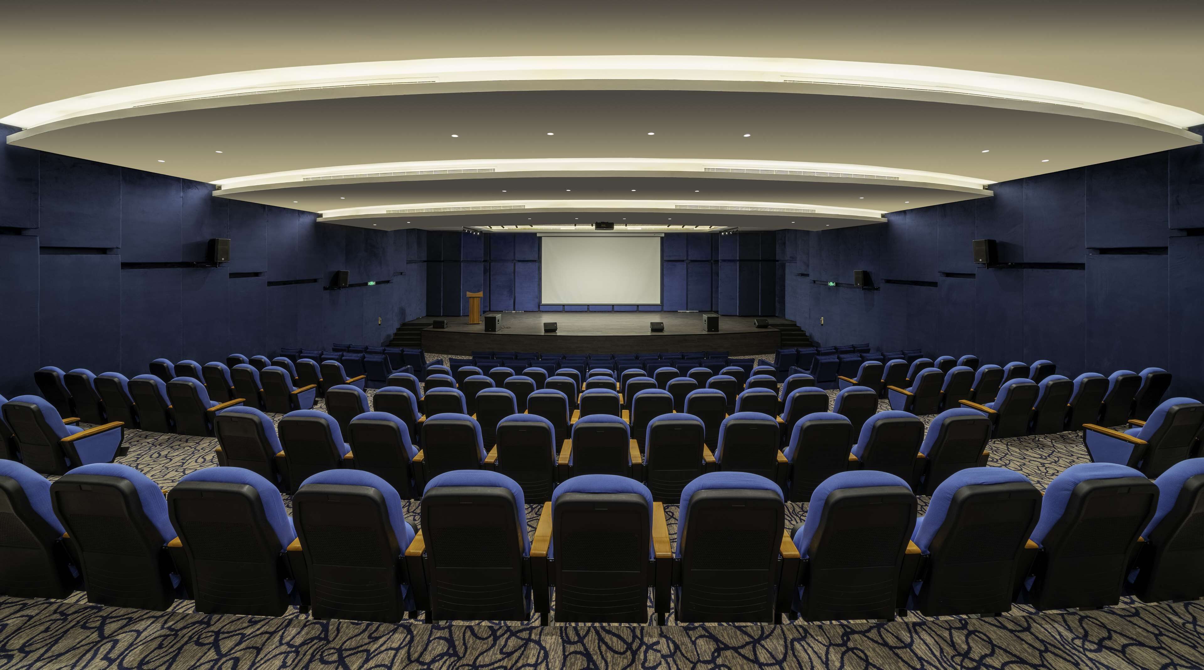 A large auditorium with rows of blue chairs, a stage with a screen, and a carpeted floor.