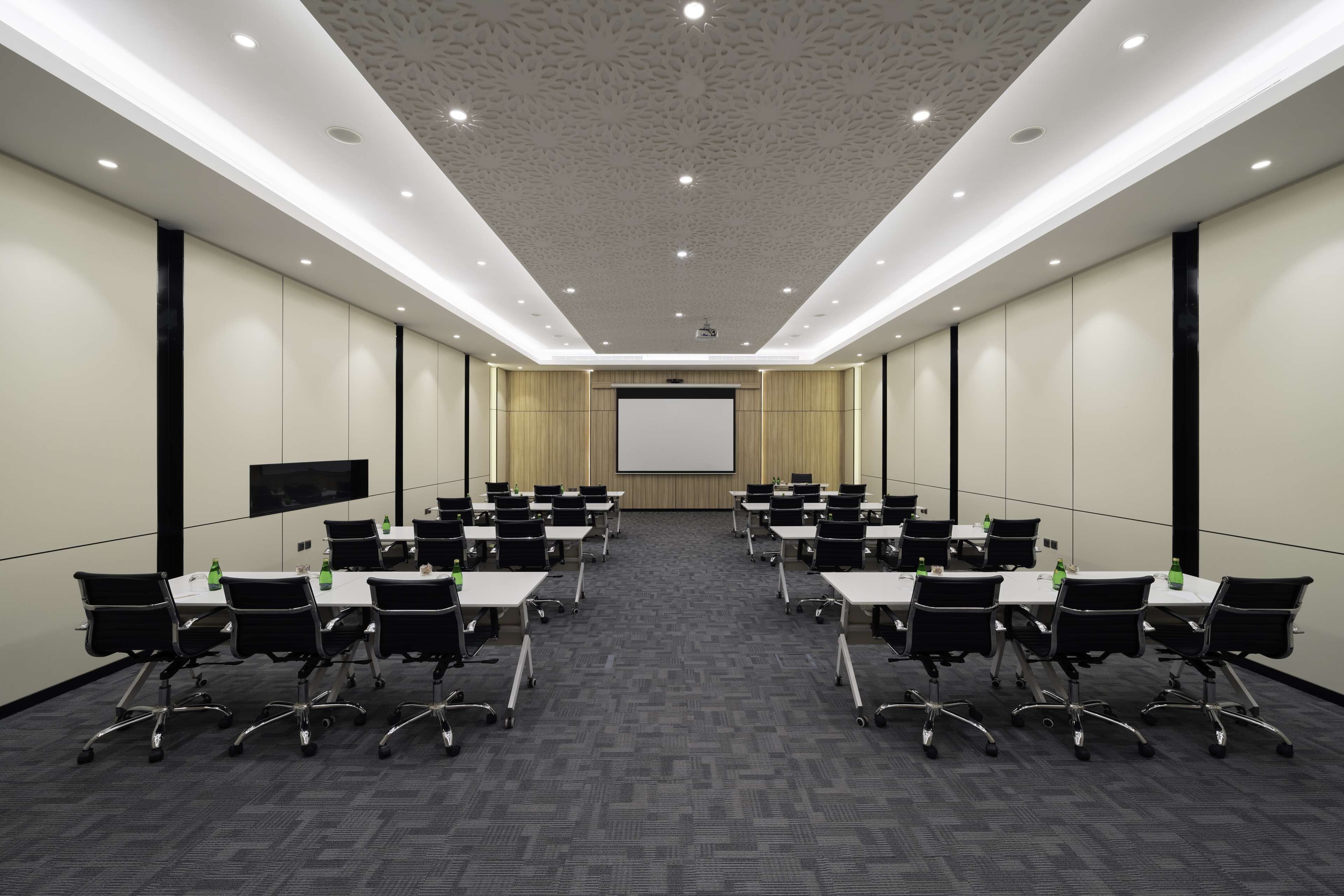 A conference room with a long table, chairs, a projector screen, and a flat-screen TV on the wall.