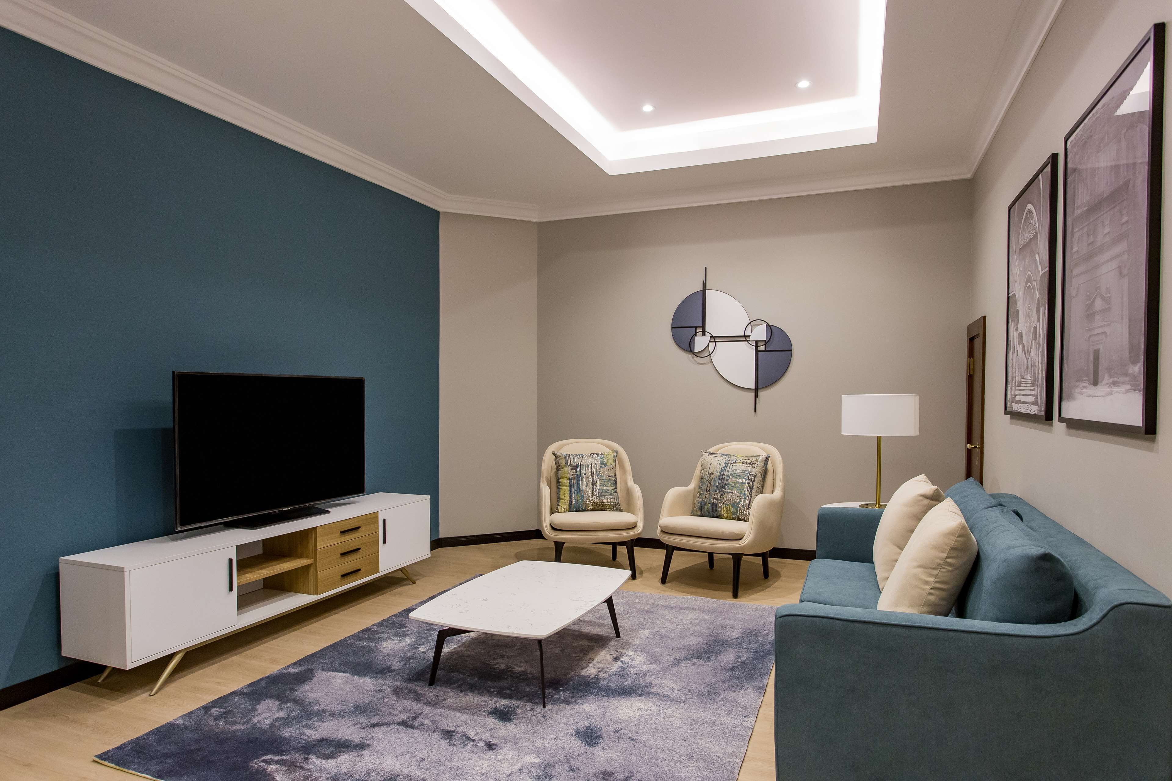 A modern living room with a flat-screen TV, a white entertainment unit, a blue sofa, two armchairs, a coffee table, and a ceiling light fixture.