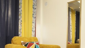 Un sillón amarillo con un cojín multicolores, un otoman turquesa y un espejo en la pared.