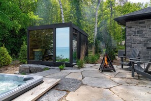Une terrasse extérieure moderne avec un sol en pierre, un jacuzzi, un espace salon clos par des baies vitrées offrant une vue dégagée et un foyer.