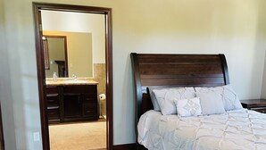A bedroom with a wooden headboard, a white bedspread, and a nightstand. There is a wooden floor and a doorway leading to a bathroom with a sink and mirror. - Luxury Lakeside/Trailside Living (Hancock)