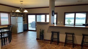 A kitchen with a refrigerator, a dining table with chairs, a bar area with stools, and large windows overlooking a body of water. - Luxury Lakeside/Trailside Living (Hancock)
