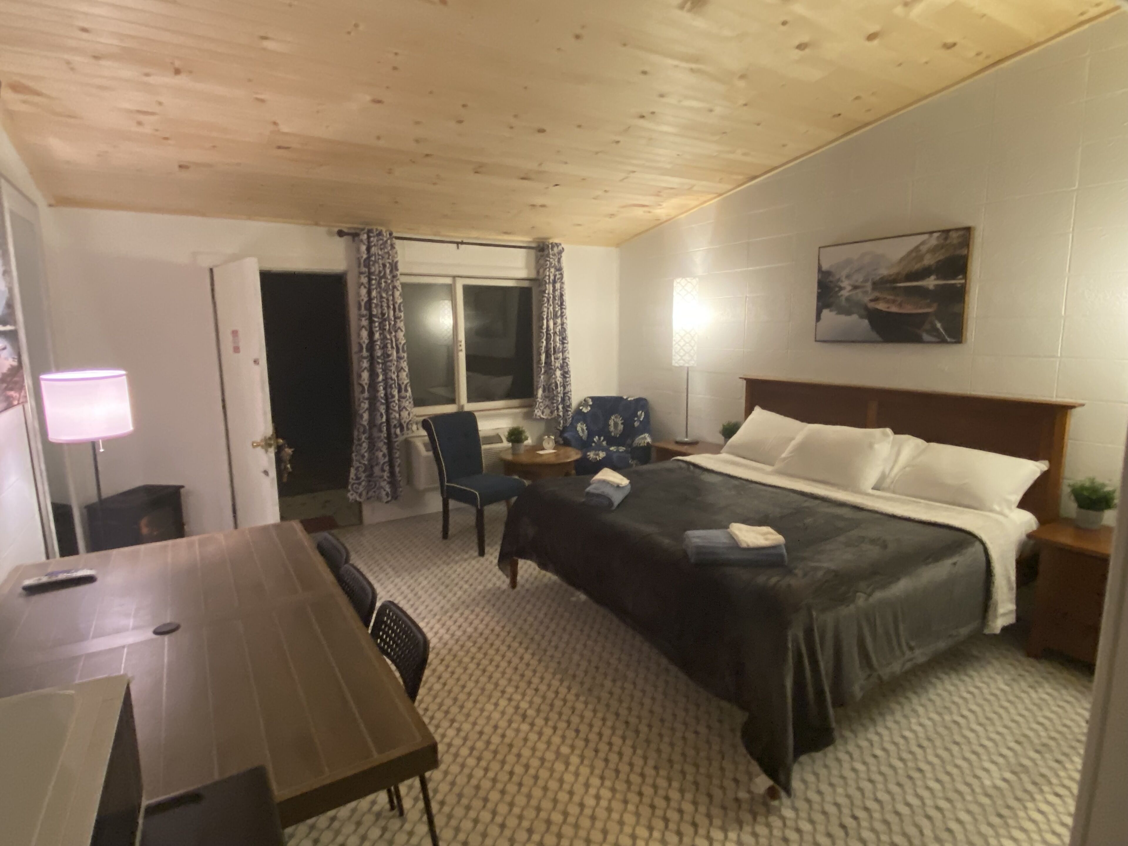 A hotel room with a wooden ceiling, a bed with white linens, a desk with a chair, a window with patterned curtains, and a painting on the wall.