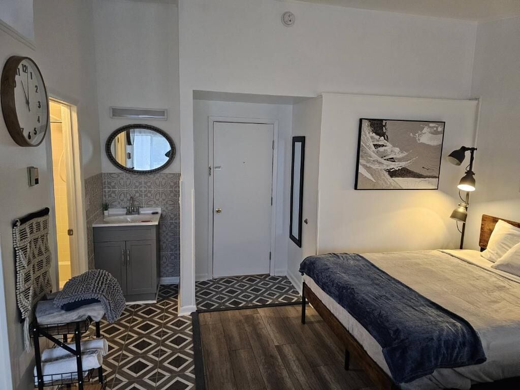 A bedroom with a bed, a bedside lamp, a wall clock, a mirror, and a door leading to a bathroom.