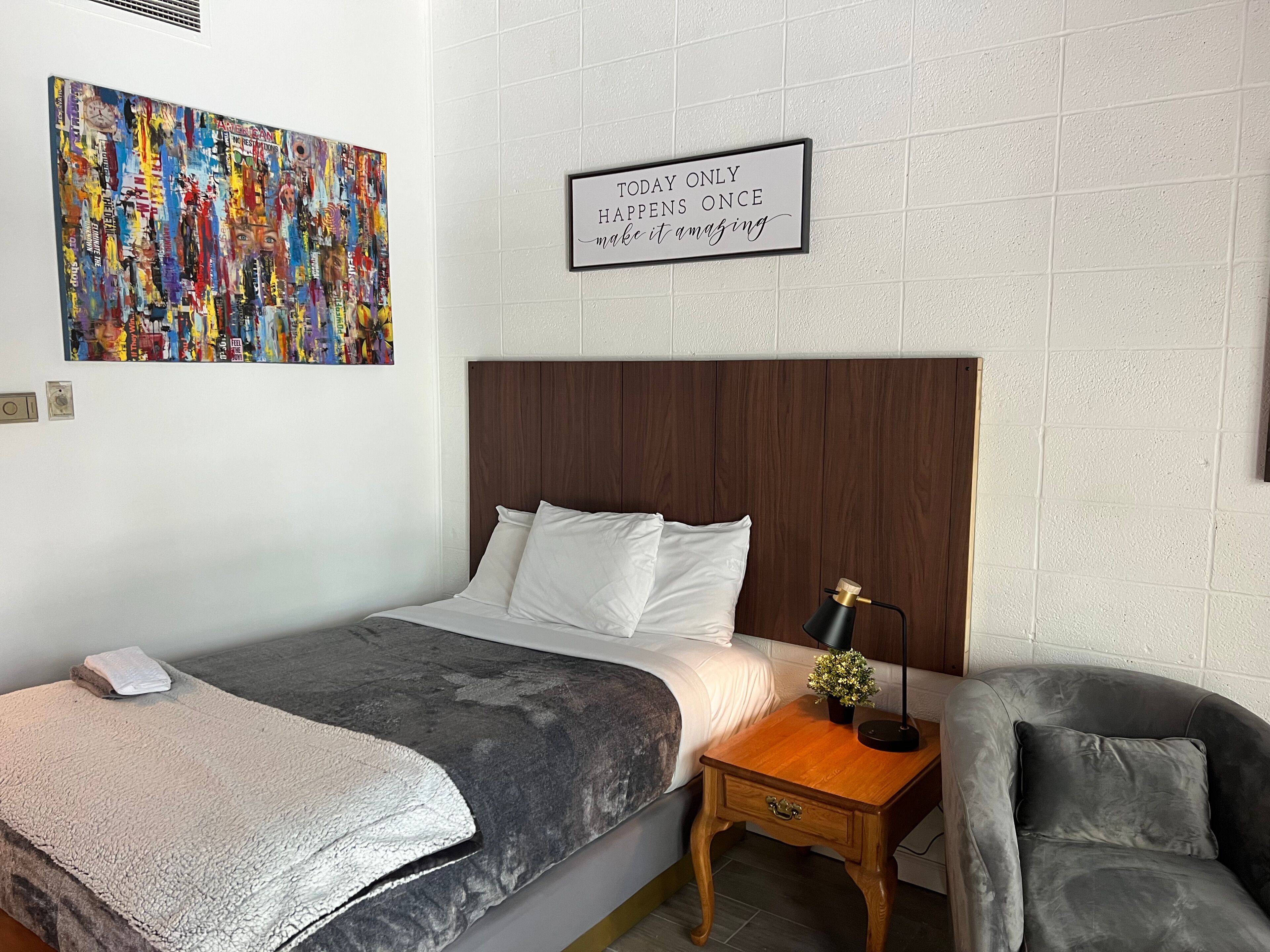 A neatly made bed with white linens, a gray textured blanket, a wooden nightstand with a lamp, and a gray armchair. A motivational quote is displayed on the wall.