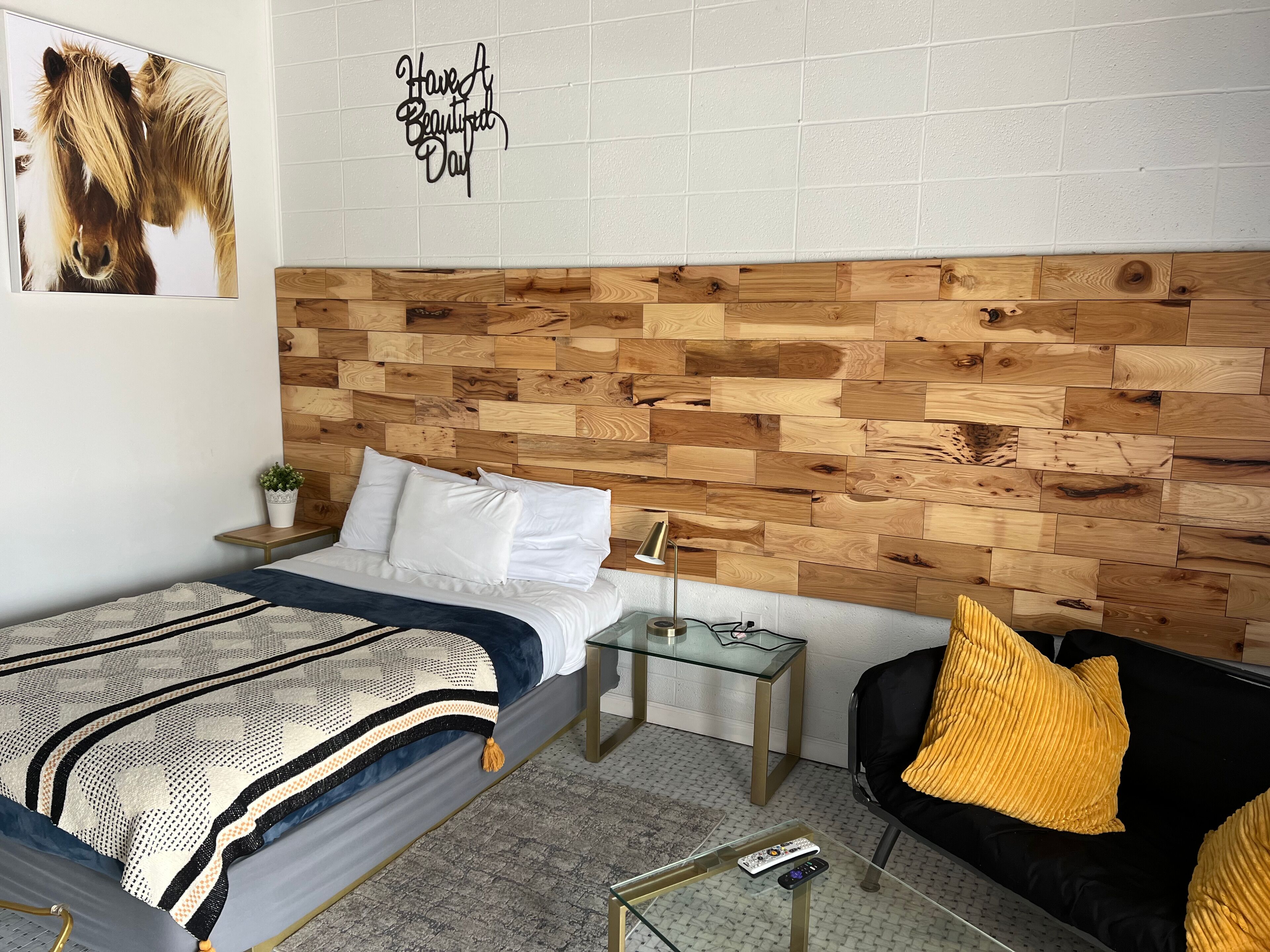 A bedroom with a wooden headboard, a bed with white bedding, a bedside table with a lamp, a small plant, and a wall with a motivational quote.