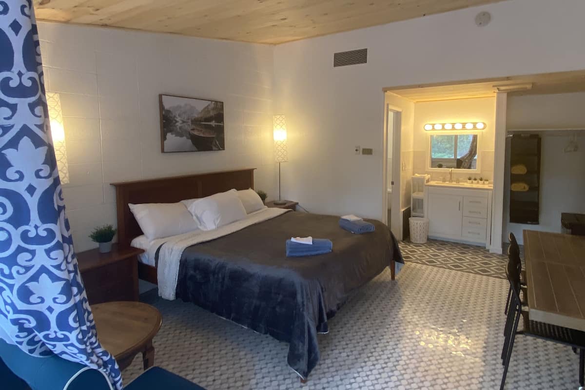 A bedroom with a bed, bedside table, lamp, and a bathroom with a sink and mirror.