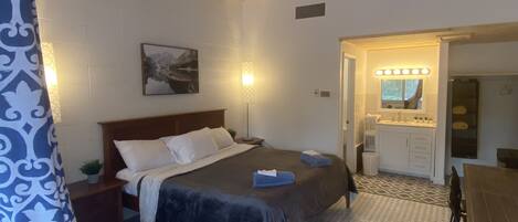 A bedroom with a bed, bedside table, lamp, and a bathroom with a sink and mirror.