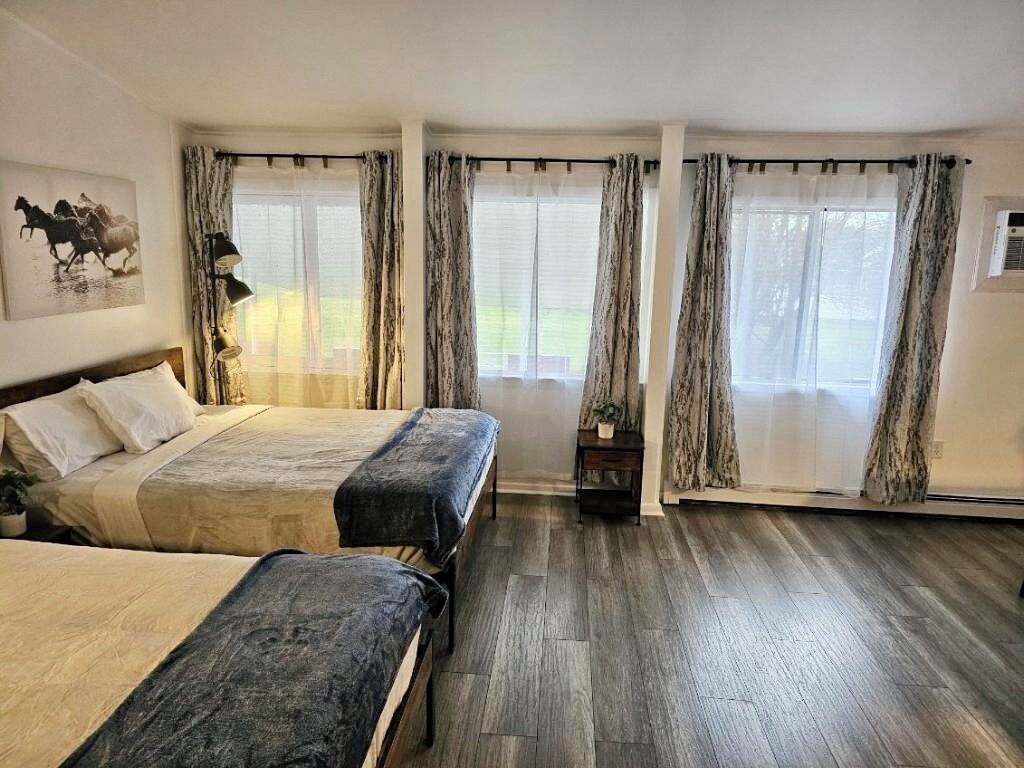 A bedroom with two beds, a large window with curtains, a lamp, and a painting on the wall.