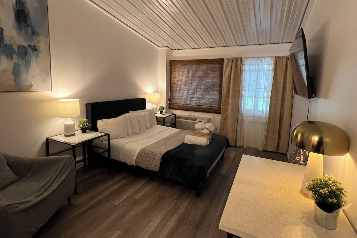 A hotel room with a bed, bedside tables, a TV, a desk, a chair, and a window with curtains.