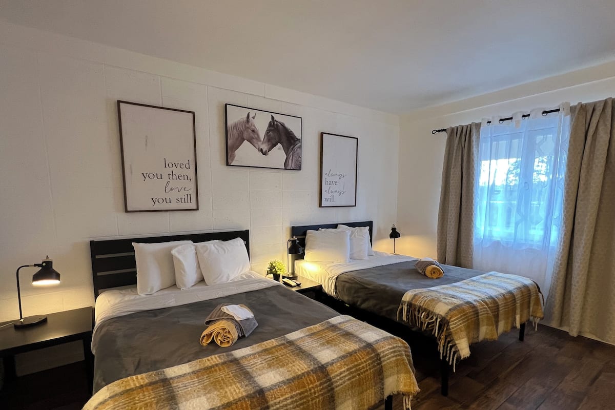 A hotel room with two beds, each with a plaid blanket and a rolled towel. There are framed pictures on the wall, a nightstand with a lamp, and a window with curtains.