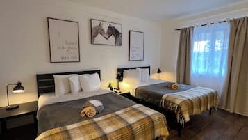 Habitación de hotel con dos camas, cada una con una manta escocesa y una toalla enrollada. En la pared hay cuadros enmarcados, una mesita de noche con lámpara, y una ventana con cortinas.