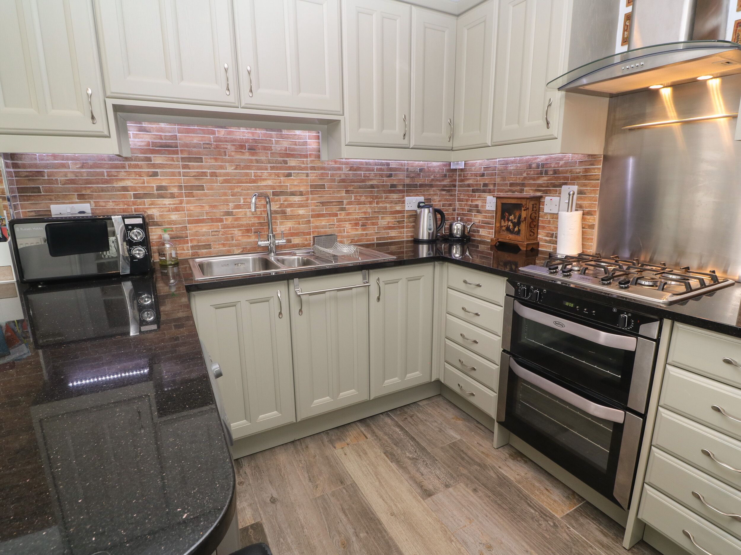 A kitchen with white cabinets, a stainless steel range hood, a double oven, a microwave, and a sink.