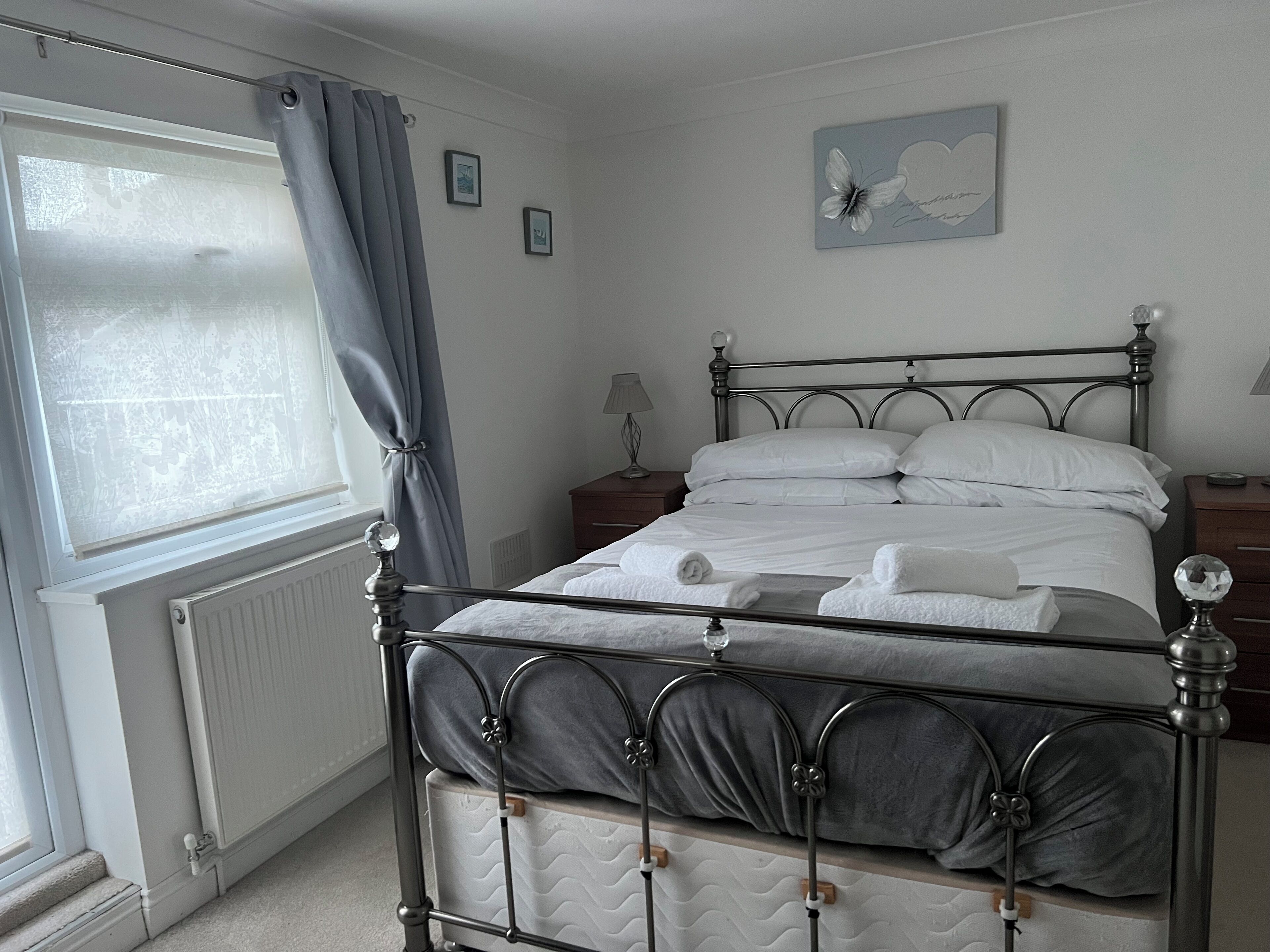 A neatly made bed with white linens and two pillows, a bedside table with a lamp, a window with a curtain, and a wall with a framed picture.