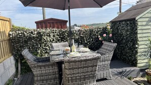 Una mesa de mimbre con cuatro sillas, un paraguas y una botella de vino, sobre una terraza de madera con un seto al fondo.