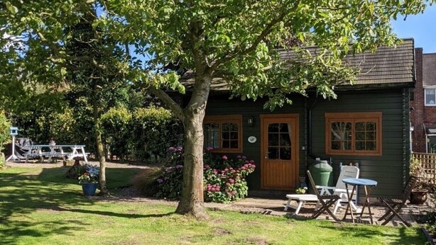 A green wooden house with a large tree in front, a small garden with flowers, and a picnic table. - Rustic Log Cabin - Dog Friendly - Sleeps 2 - Views (Shrewsbury)