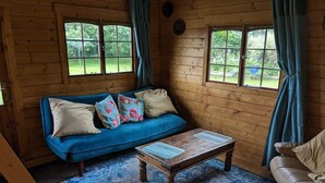 A cozy room with a blue sofa, a wooden coffee table, and a leather chair. There are two windows with curtains, and a wooden floor. - Rustic Log Cabin - Dog Friendly - Sleeps 2 - Views (Shrewsbury)