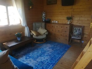 A cozy room with a wooden floor, a blue patterned rug, a cushioned chair, a small table, and a decorative chair with a horse painting. - Rustic Log Cabin - Dog Friendly - Sleeps 2 - Views (Shrewsbury)